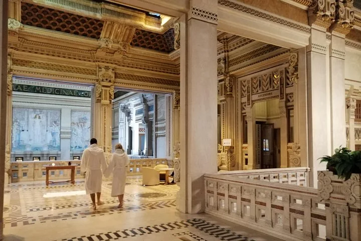 Two guests in white bathrobes walking across an ornate marble atrium with painted panels and carved columns